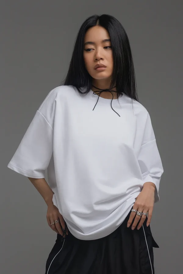 A young East Asian woman wearing an oversized plain white T-shirt, black skirt with white piping, and multiple silver rings, standing against a gray studio background with a confident expression.
